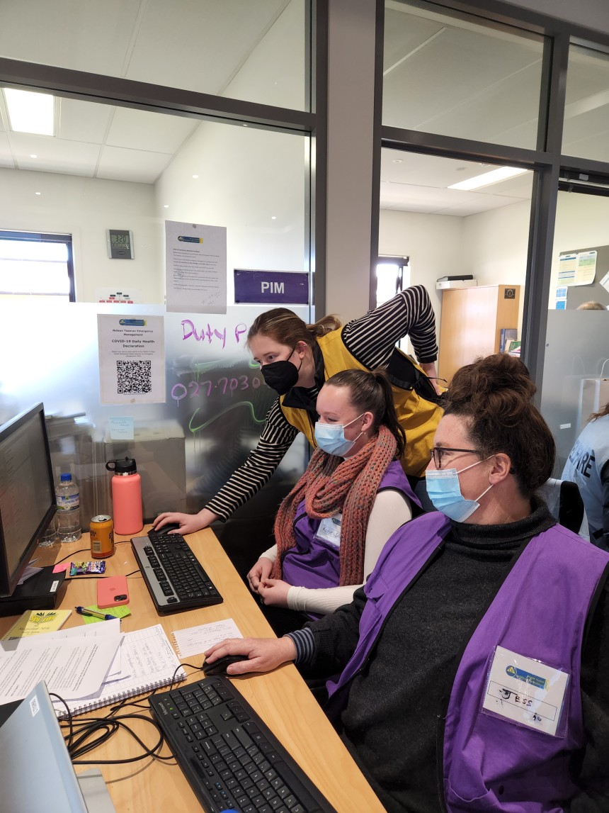 Photo: Two woman wearing face masks are in an emergency operations centre at a desk in front of computers. Another woman is standing and using a computer mouse to show them something. 