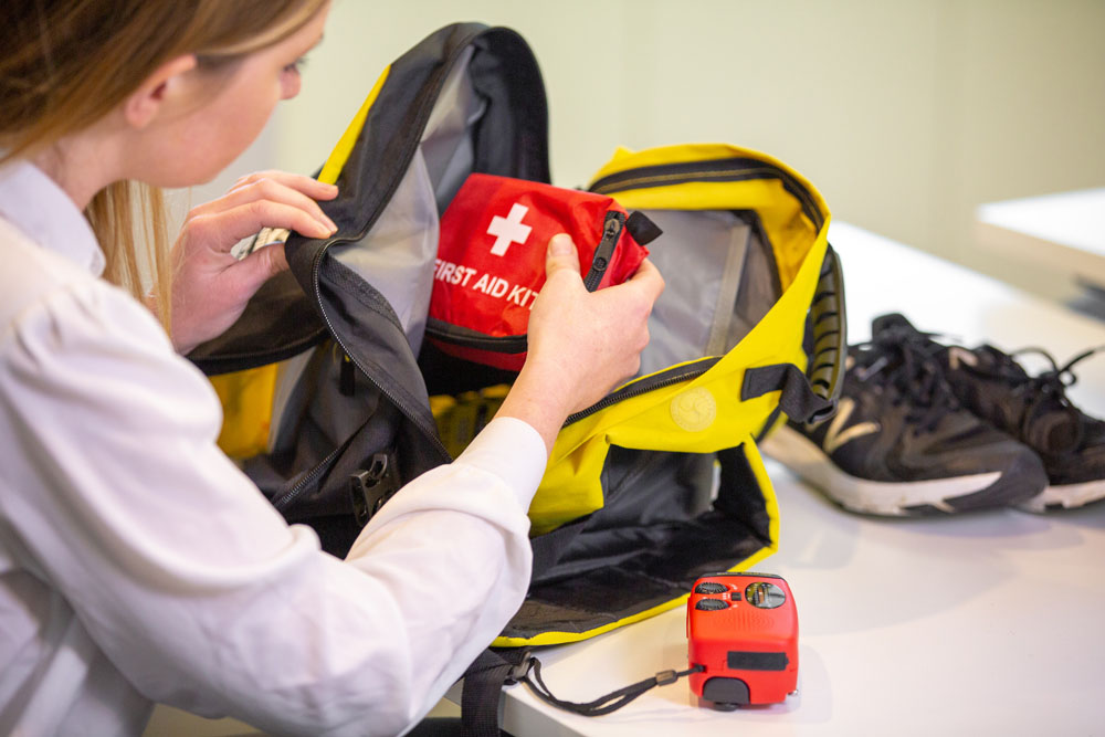 Woman placing first aid kit inside bag. On the table is a radio/torch and a pair of running shoes