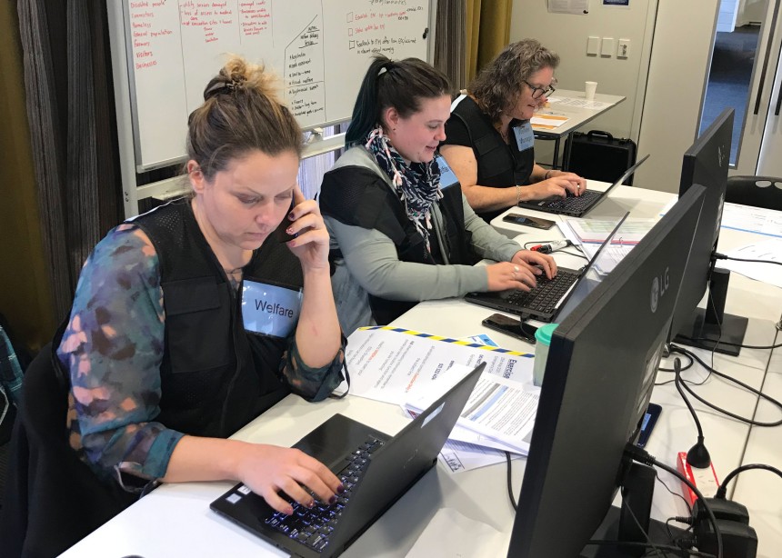 Photo: 3 women are in front of laptops in an emergency operations centre. They are wearing vests with pockets that say welfare. There is a whiteboard in the background with information written on it